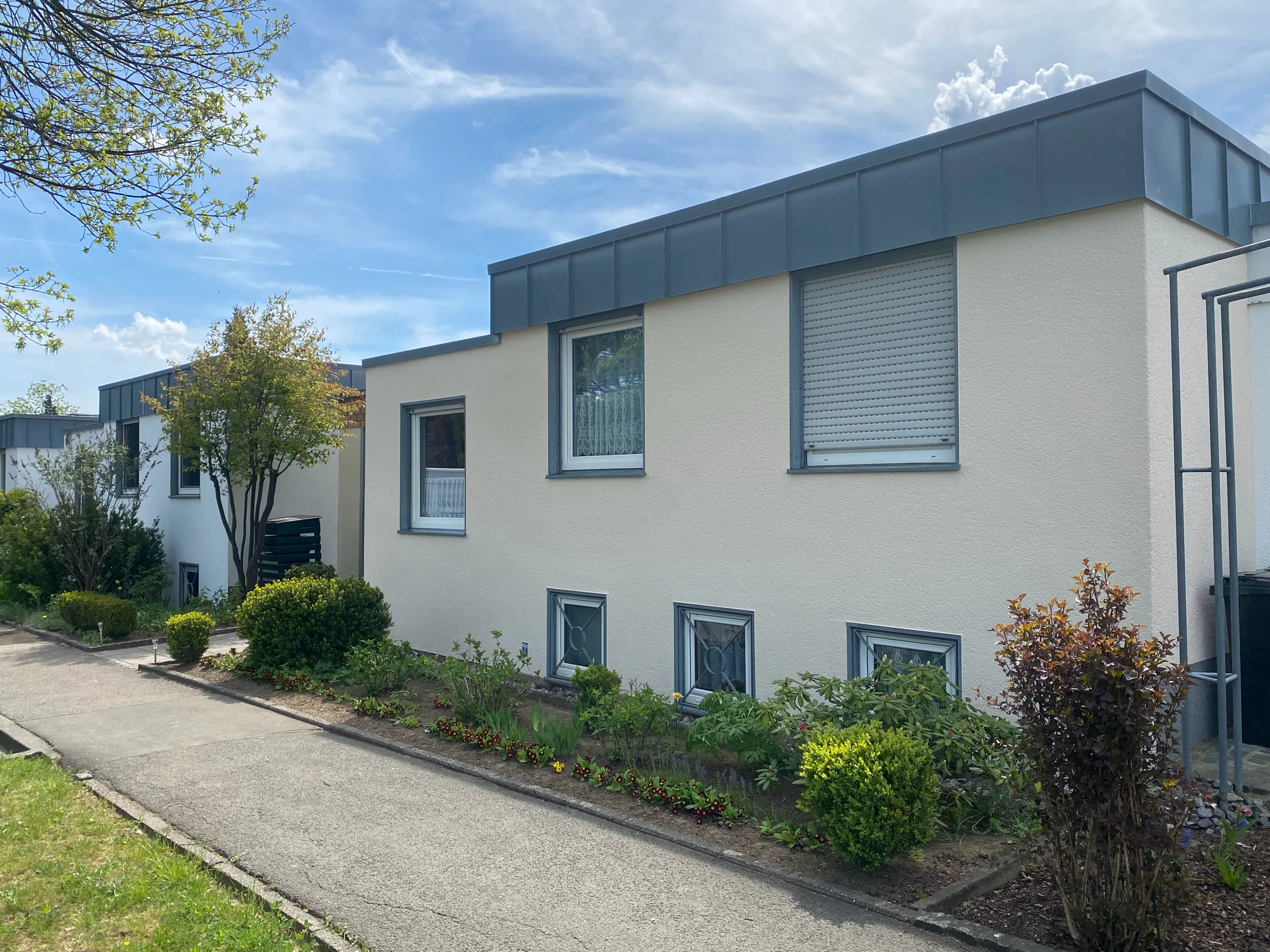 Heller Bungalow mit Flachdach und frisch gestrichener cremefarbener Fassade, davor ein blühender Vorgarten